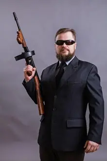 Mafia Prints: Mature bearded man in sunglasses dressed in suit with tommy gun on grey background