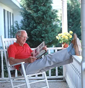 Porch Prints: mature man relaxing on rocking chair on wooden porch looking at photo album