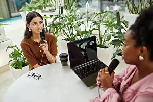 Medium shot of young businesswoman live streaming podcast with female guest sitting at cafe table equipped with laptop