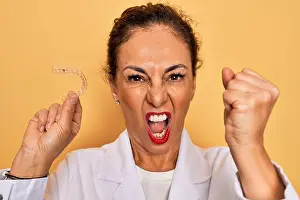 Dental Prints: Middle age senior dentist woman holding clear aligner for teeth correction annoyed and frustrated shouting with anger