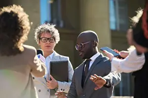 Middle Aged Prints: Middle aged Caucasian woman and young Black man standing outdoors surrounded by journalists holding microphones