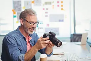 Dslr Prints: Middle-aged man in denim shirt inspecting DSLR camera at office desk with coffee cup and notebook