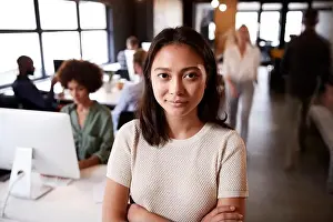 Arms Crossed Prints: Millennial Asian female creative standing in a busy casual office, smiling to camera