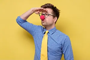 Trouble Prints: Millennial man in clown clothes with red nose and glasses looking aside far away on yellow wall