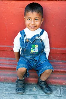 Eyed Prints: mischievous bright eyed smiling little Mexican boy sits outside against deep red wall on a street in Oaxaca de Juarez