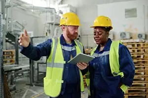 Multicultural Prints: Mixed-race female factory worker in hardhat and uniform talking to male colleague using tablet in workshop