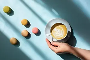Hot Drink Prints: Morning coffee in a woman's hand with delicious macaroons scattered around in the sunlight