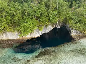 The mouth of a dark, water-filled cave opens on the edge of a forest-covered limestone island near Fak Fak, Indonesia. Limestone often harbors caves