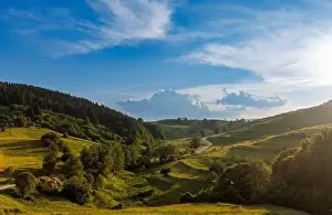A narrow automobile paved intermountain road passes and meanders among low hills covered with spruce forests