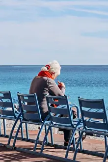French Riviera Prints: Nice, France - November 7, 2022: Old lady sitting on the famous blue chairs in Nice on the Promenade des Anglais