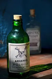 Deadly Prints: old green glass bottle labelled with arsenic poison and skull and bones stands on a shelf