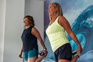 Flexibility Prints: Two older women in sportswear are smiling as they stretch their quadriceps muscles during a training routine in the gym
