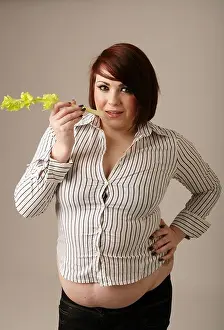 Overweight woman in a tight fitting shirt standing holding a stick of celery