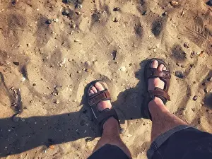 Mans Prints: A pair of male feet wearing sandals on a sandy beach
