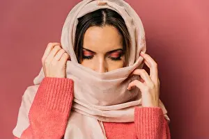 Headwear Prints: Peaceful arab female with makeup covering face with long light headscarf while standing on pink background with closed