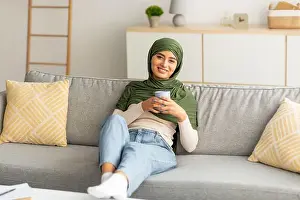 Lockdown Prints: Peaceful weekend. Positive young Arab lady in hijab relaxing on sofa with cozy hot drink at home