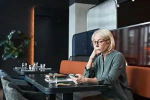 Professional Occupation Prints: Serious pensive attractive senior lady with blond hair sitting at table in modern cafe in black color