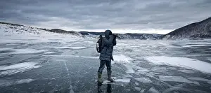 Rippled Prints: Person in winter gear explores and photographs the icy surface of a winter lake surrounded by snowy hills