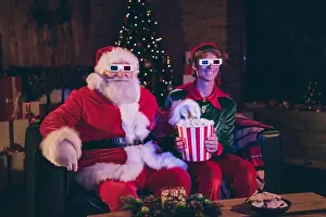 Holiday Home Prints: Photo portrait of santa claus and elf eating popcorn watching movie on sofa in 3d glasses
