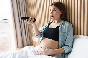Shocked Prints: Photo of shocked pregnant woman using remote control while watching tv in bed at sunny room