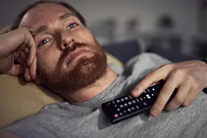 Exhausted Prints: Portrait of bearded adult man watching TV at night while lying on couh in dark room and switching channels