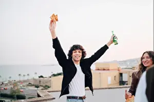 Pizza Prints: Portrait of carefree man holding beer bottle and pizza slice at rooftop party
