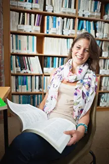 Bookshelf Prints: Portrait of a happy female student sitting on chair and with a book in the library