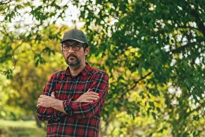 Arms Crossed Prints: Portrait of male farmer with arms crossed wearing plaid shirt and trucker's hat posing in walnut orchard