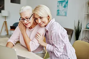 Assist Prints: Portrait of smiling young woman embracing senior mother while looking at laptop screen together in cozy home interior
