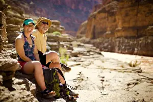 Weekend Activities Prints: Portrait of a smiling young woman and mature man sitting side by side on a rock near a riverbank inside a canyon