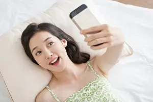 Korean Ethnicity Prints: Portrait of young smiling woman lying down on bed and taking picture of herself with smartphone