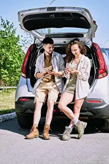 Couple Relationship Prints: Positive active young hikers in boots leaning on car trunk and discussing hiking route using phone apps