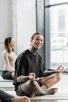 Meditate Prints: Positive tattooed man looking at camera while doing gyan mudra in yoga studio, stock image
