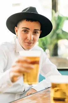 Goblet Prints: Positive tomboy with tattoos wearing hat and white shirt drinking beer from glass in cafe