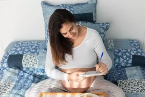 Prenatal Prints: From above of pregnant female sitting on bed and writing in notebook during breakfast in morning