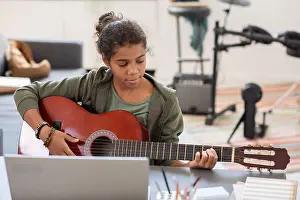 Preteen Prints: Pretty biracial girl playing acoustic guitar in front of laptop while sitting by table against drumkit in bedroom