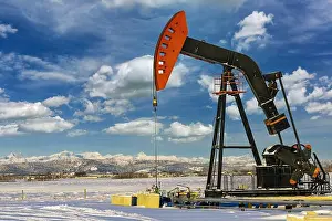 Prairies Prints: Pumpjack in a snow-covered field with dramatic clouds in a blue sky with snow-covered mountain range in the background