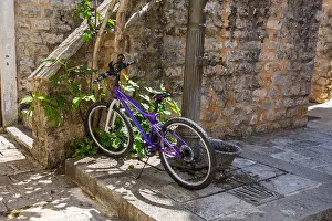 Spoke Prints: Purple bicycle leaning against old water pump in narrow stone alley
