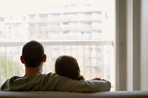 Rear view of couple sitting on sofa looking out of window at apartment building