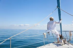 Sail Boat Prints: Rear view of a senior woman alone sitting on the front of a sail boat on a calm blue sea
