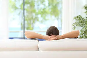 Meditating Prints: Rear view of a single man relaxed on a couch at home and looking the green background outdoors through the window