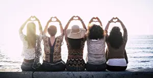 Admiration Prints: Rear view of five women sitting in a row on sea wall gesturing love by making heart shape symbol with hands