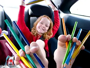 Toes Prints: Red haired girl with multi-coloured pencils between her ten toes in the back of a car