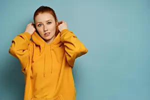 Redhead Prints: The red-haired girl stands in a yellow hoodie against a blue background, lifting the hood