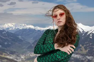 Redhead Prints: Red-haired young woman posing in green jumpsuit and glasses against the backdrop of snow-capped mountains