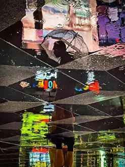 Reflection of a standing man holding an umbrella surrounded by colorful neon lights as seen in the reflection on wet