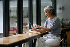 Senior Women Prints: A relaxed and happy Asian retired lady in casual clothes enjoys the app on her smartphone while sitting in a coffee