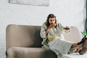 Remote Controller Prints: selective focus of Jesus in crown of thorns watching tv and eating popcorn on sofa at home