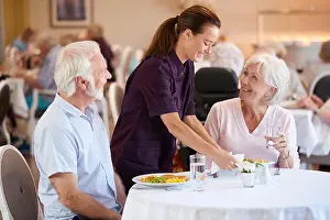Caring Prints: Senior Couple Being Served With Meal By Carer In Dining Room Of Retirement Home