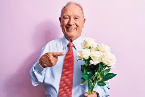 Optimistic Prints: Senior grey-haired businessman wearing tie holding bouquet of flowers over pink background smiling happy pointing with